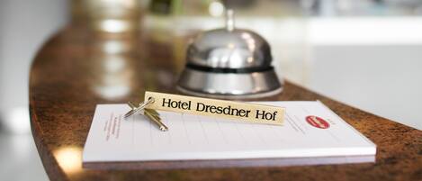 Mostrador de recepción de hotel con una tarjeta de identificación que dice "Hotel Dresden Hof", una campana y un bolígrafo sobre un bloc de notas.