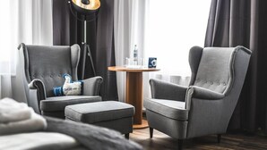 Una habitación de hotel moderna con dos sillones grises, una pequeña mesa redonda de madera con una botella y vasos, y una lámpara de pie.