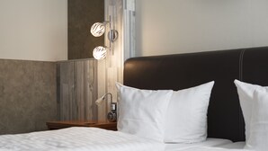 Una cama bien hecha con sábanas blancas, una mesita de noche de madera con una lámpara y un aplique de pared.