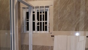 A bathroom with a glass shower enclosure, a toilet, a sink, and a window with bars. - Dream Vacation ST Catherine Jamaica - Guest Suites for Rent in Spanish Town (Spanish Town)