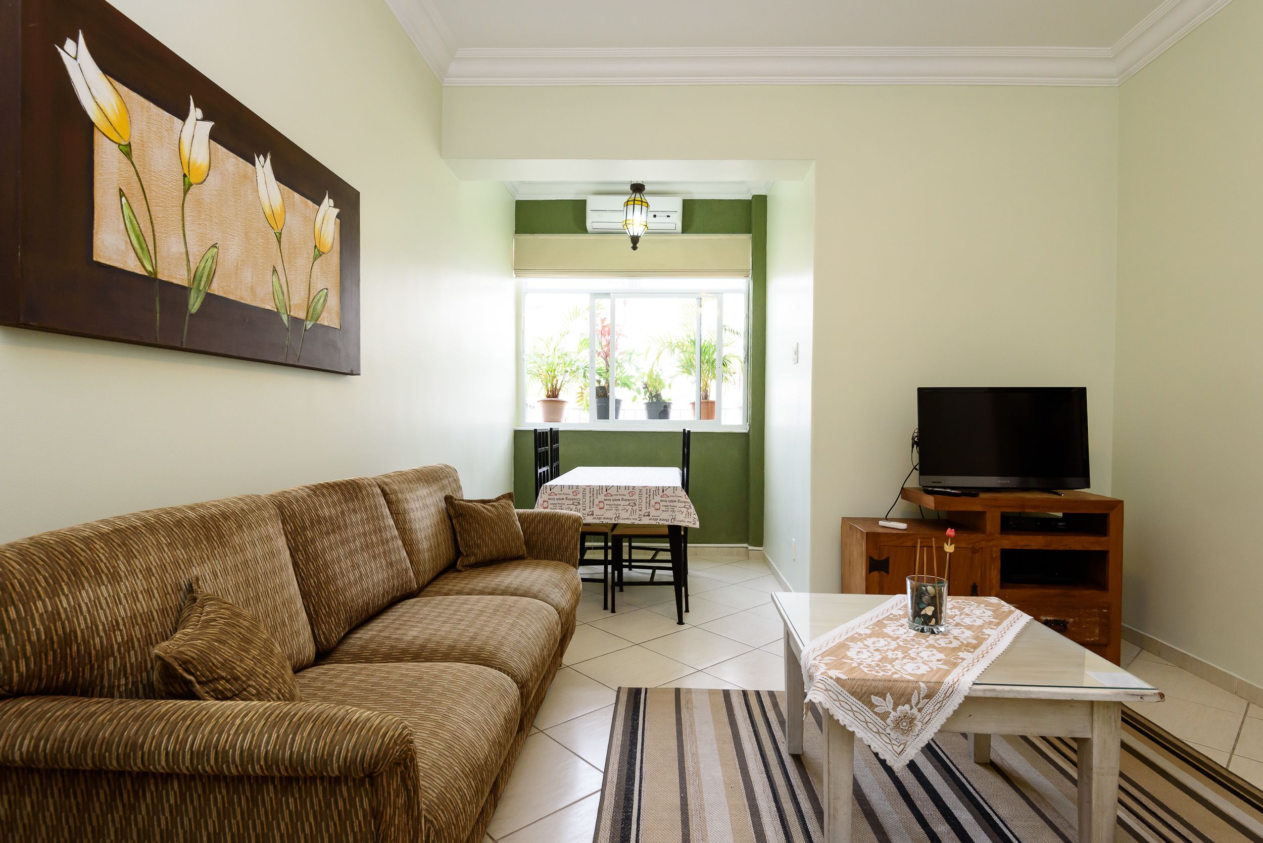 A living room with a brown sofa, a television on a wooden stand, a dining table with chairs, and a painting on the wall.