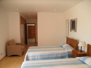 Un dormitorio con dos camas, una silla, una mesita de noche y una puerta que da a otra habitación.