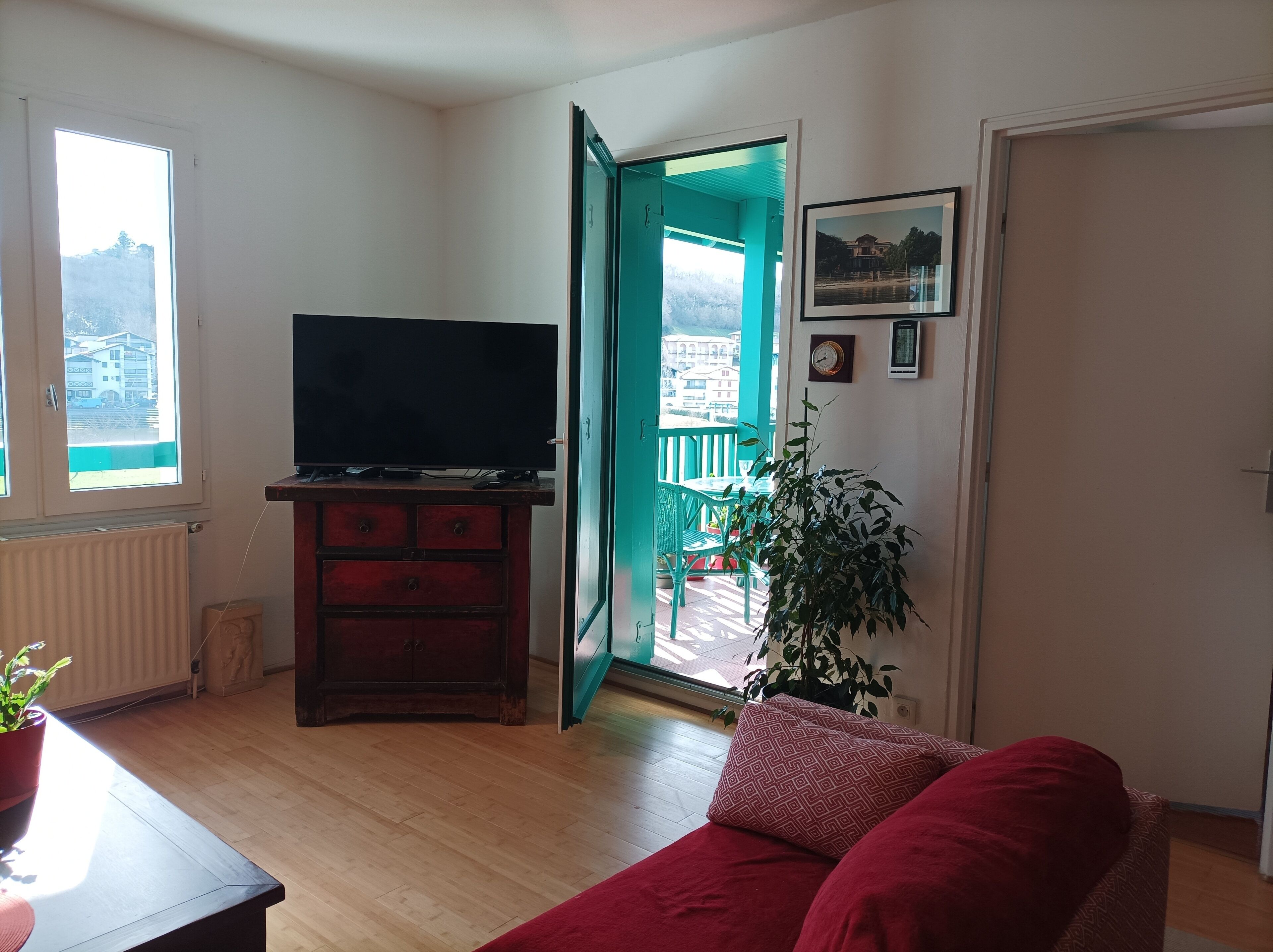 Un salon avec un téléviseur posé sur un meuble en bois, un canapé rouge, une plante en pot et un balcon offrant une vue sur les immebles.