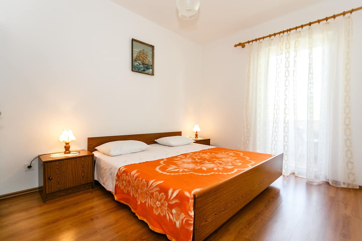 Un dormitorio con una cama de madera, dos lámparas de noche, una mesita de noche y una ventana con cortinas traslúcidas.