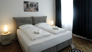 Een net opgemaakt tweepersoonsbed met witte lakens en handdoeken, twee bedlampen, een ingelijste afbeelding aan de muur en een donker gordijn voor het raam.