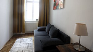 Een moderne woonkamer met een donkere bank, een houten salontafel en een lamp. Er is een raam met gordijnen zichtbaar, en er hangt een schilderij aan de muur.