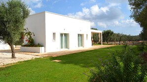A modern white building with a flat roof, a covered patio area, and a well-maintained lawn with various plants. - Casa Kairos (Ostuni)