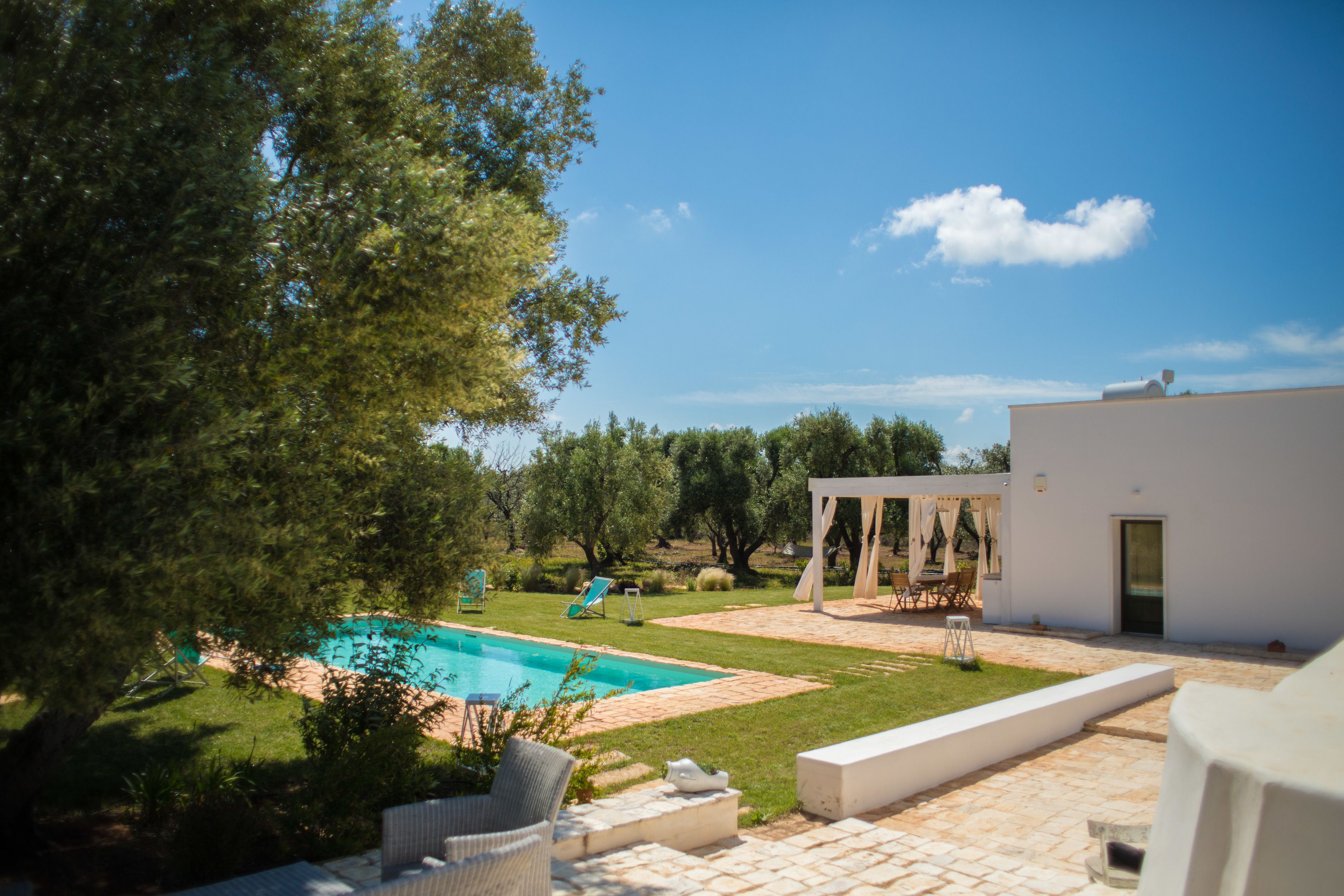 Area piscina con un edificio bianco, arredi da esterno e una pergola.