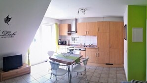 Een kleine keuken met houten kastjes, een eettafel met stoelen, een televisie op een standaard en een raam voor natuurlijk licht.