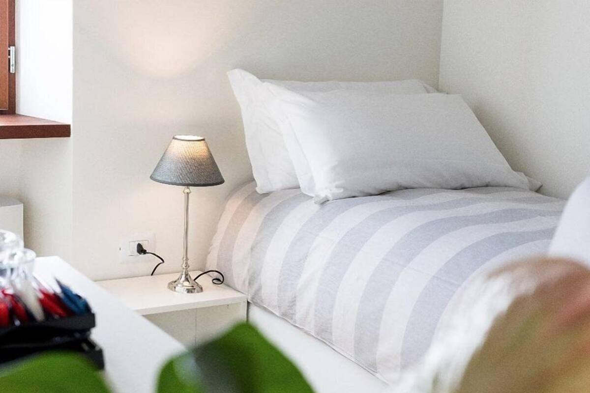 A neatly made bed with striped bedding, a bedside lamp, and a power outlet on the wall.