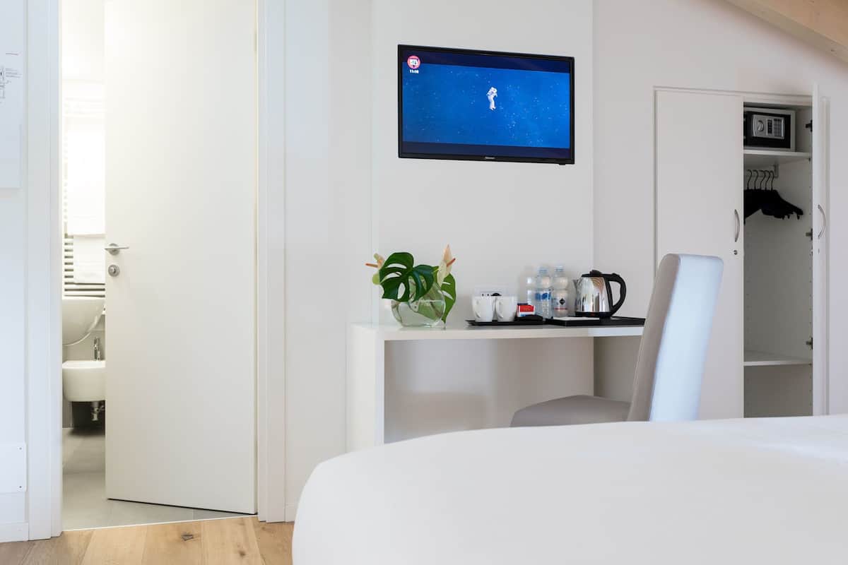 A modern hotel room with a flat-screen TV mounted on the wall, a small desk with a vase of flowers, a chair, and a wardrobe with a hanging rack.