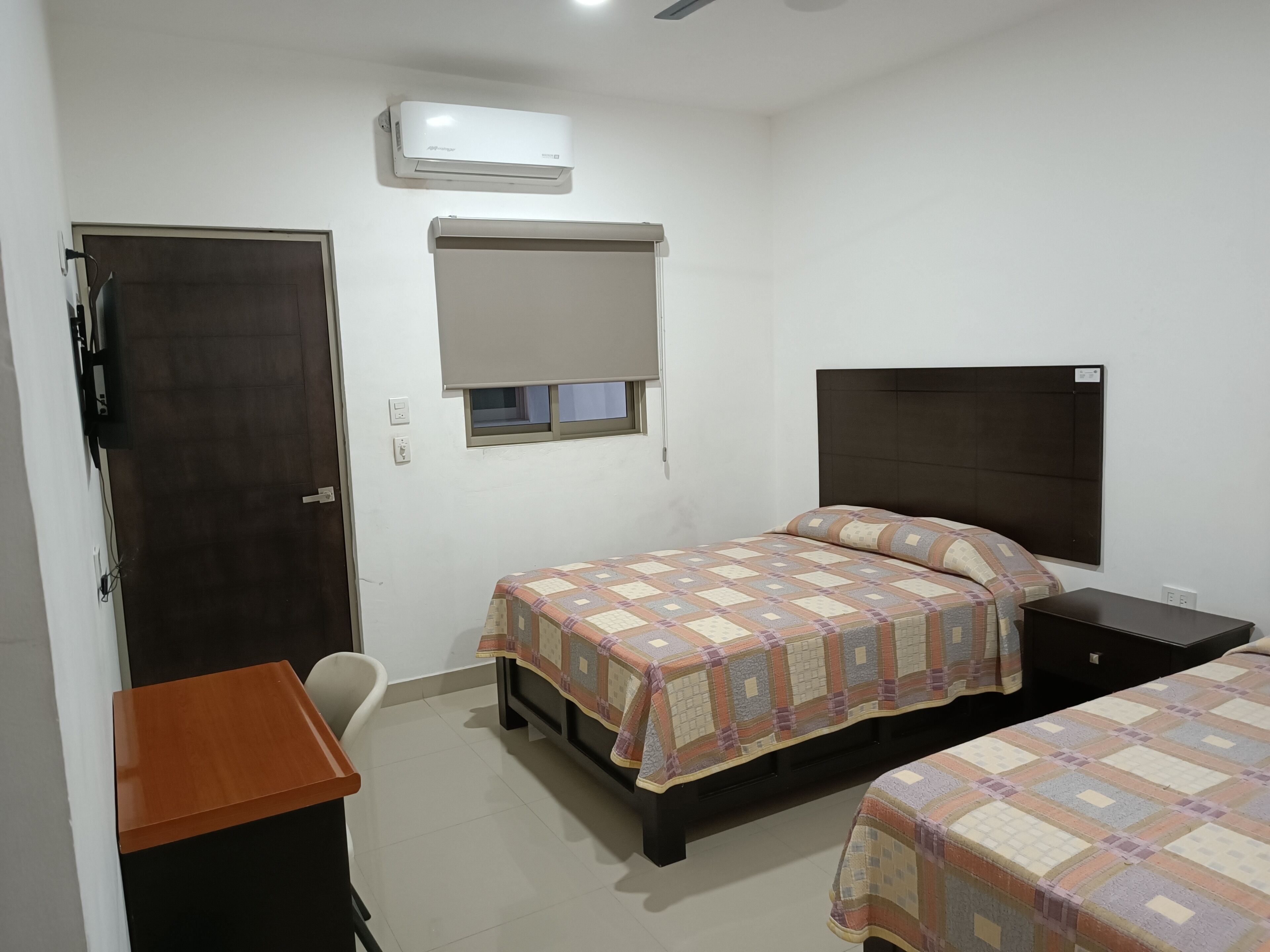 Un dormitorio con cama, escritorio, silla, ventana con persiana y equipo de aire acondicionado.