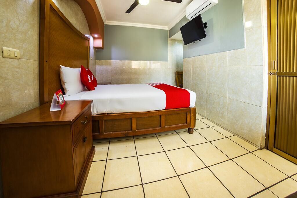 A hotel room with a bed, wooden headboard, a nightstand, a TV mounted on the wall, and a tiled floor.