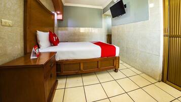 A hotel room with a bed, wooden headboard, a nightstand, a TV mounted on the wall, and a tiled floor.