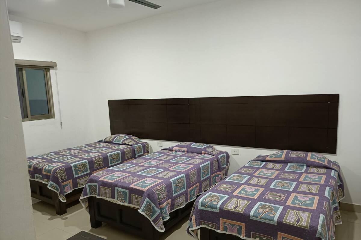 A room with three single beds, each with a patterned bedspread, a dark headboard, and a small bedside table.
