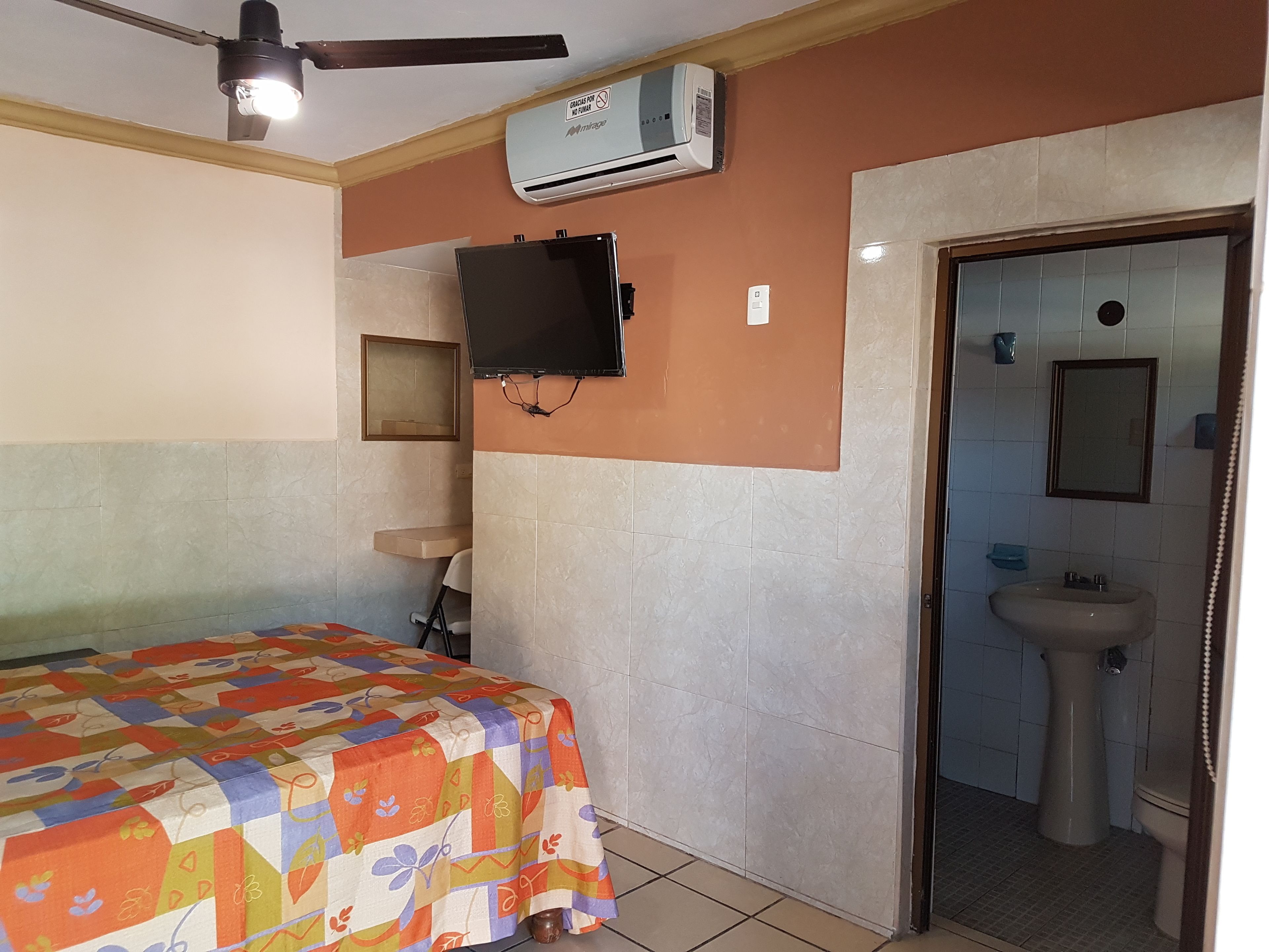 A hotel room with a bed, a television mounted on the wall, an air conditioning unit, a sink, and a mirror in the bathroom.