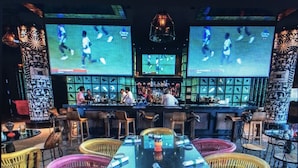 Angolo bar con diversi schermi che mostrano eventi sportivi, banconi e sedie in vimini.