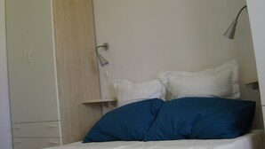 Un dormitorio con cama, almohadas blancas, una almohada azul, una lámpara de noche y un armario.