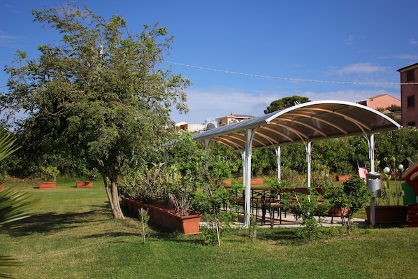 A covered outdoor seating area with tables and chairs, surrounded by potted plants and trees.