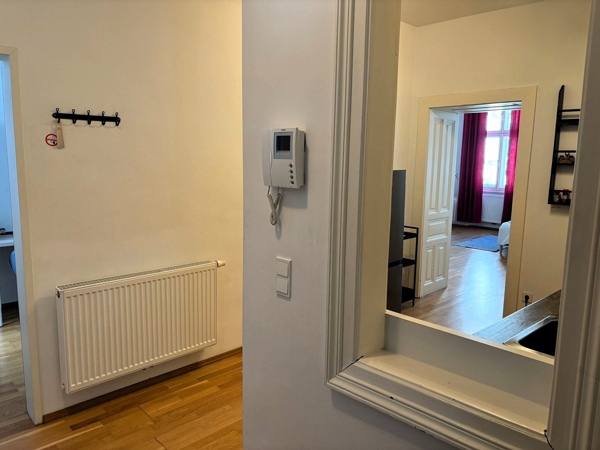 En gang med en hvid radiator, et intercom på væggen og et spejl der reflekterer et rum med en seng og et vindue med røde gardiner.