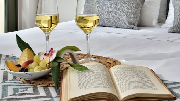 Una cama bien hecha con almohadas, una mesita de noche con dos copas de vino blanco, un frutero y un libro abierto.