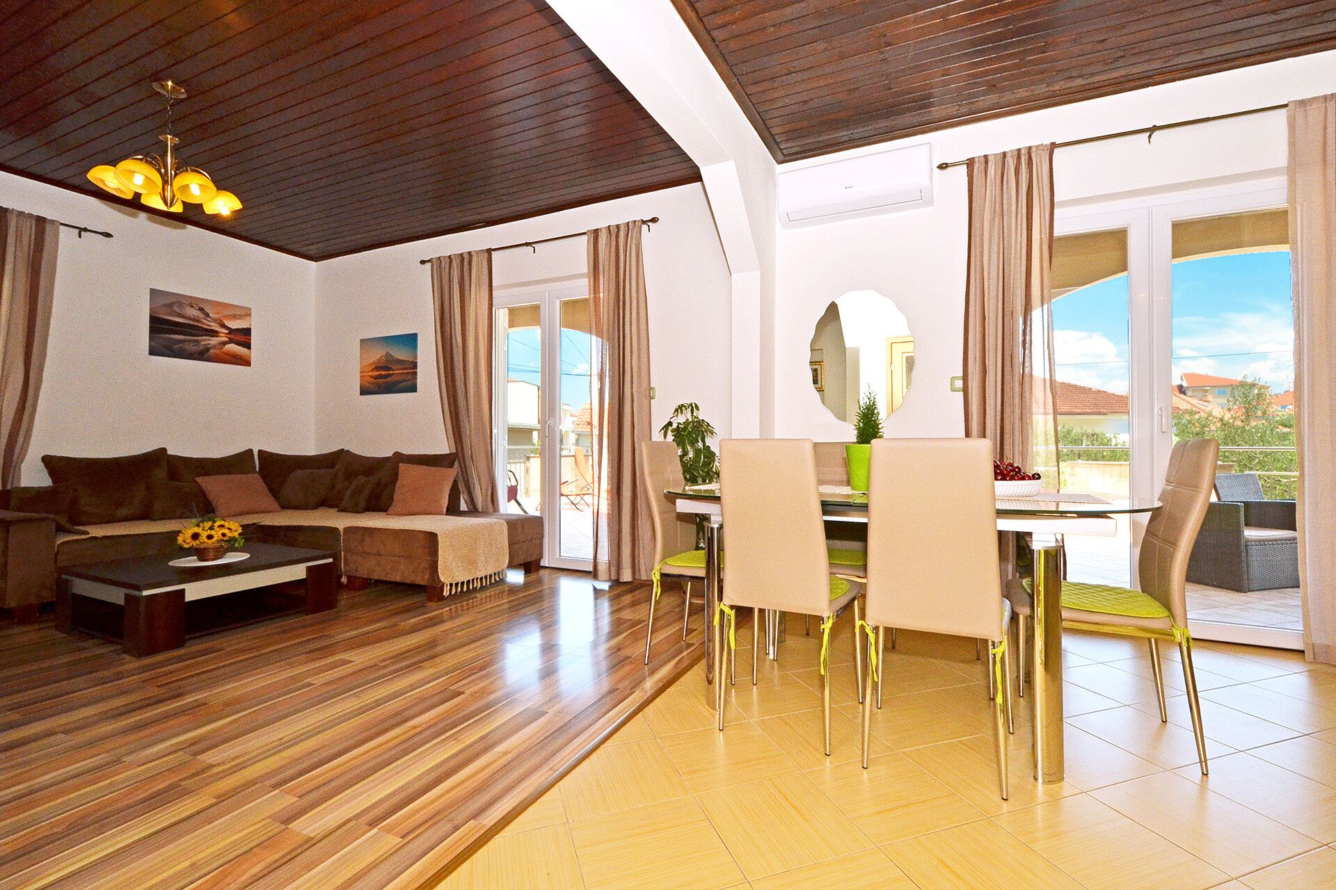 A modern living room with a wooden ceiling, a sofa, a coffee table with a flower vase, a dining area with chairs and a table, and a large window with a view of the outdoors.