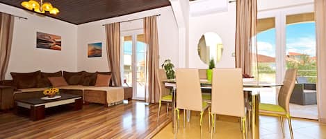 A modern living room with a wooden ceiling, a sofa, a coffee table with a flower vase, a dining area with chairs and a table, and a large window with a view of the outdoors.