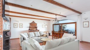 A spacious living room with a sectional sofa, a wooden dining table with chairs, a fireplace, and a large window offering a view. - Our sea view house offers space on 400 square meters for 2-10 people (Magomadas (OR))