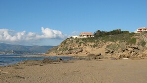 A coastal beach with a sandy shore, rocky outcrops, and a building with a red roof on a hillside. - Our sea view house offers space on 400 square meters for 2-10 people (Magomadas (OR))