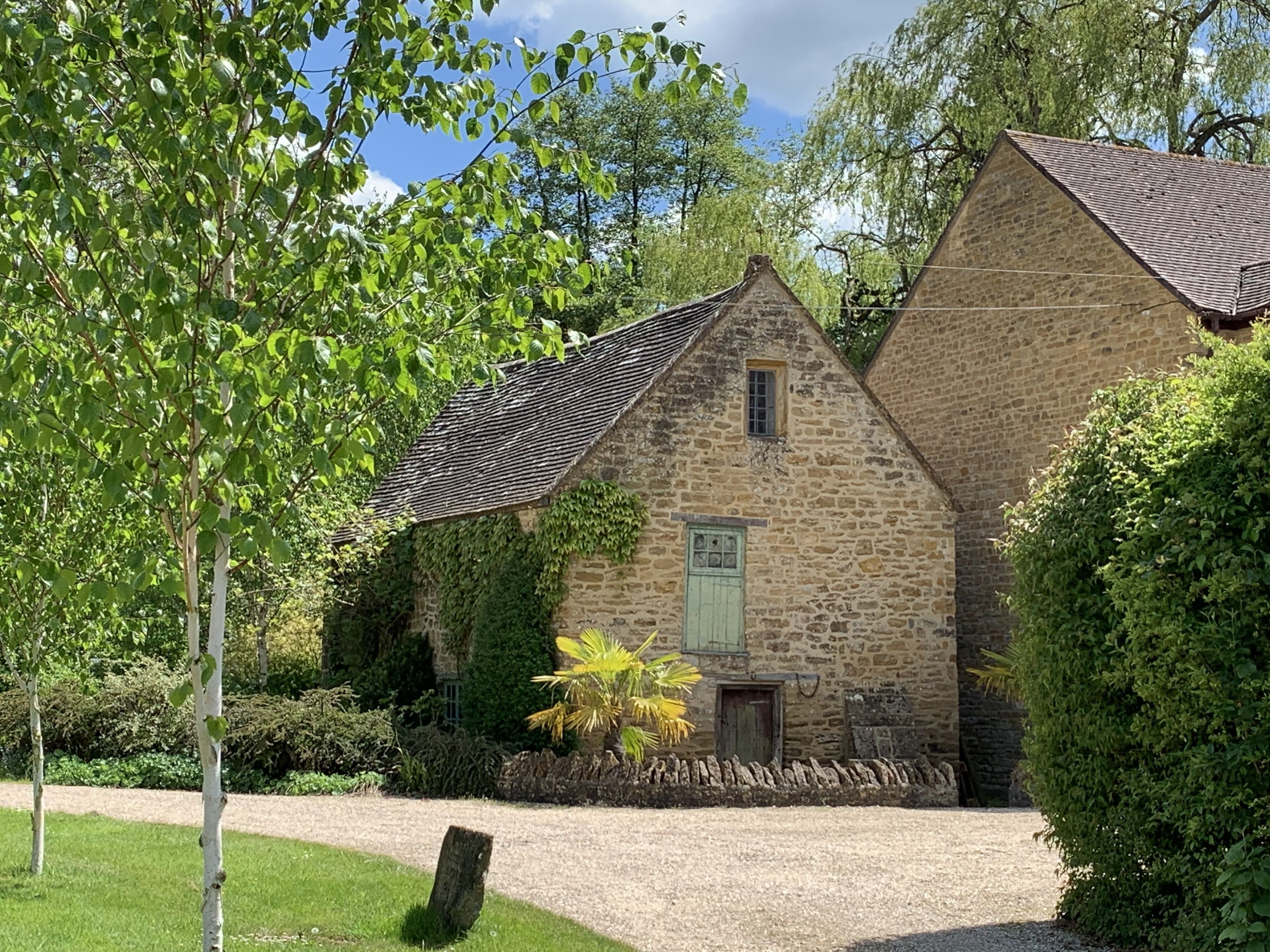 A stone building with a sloped roof, surrounded by greenery and a gravel path.