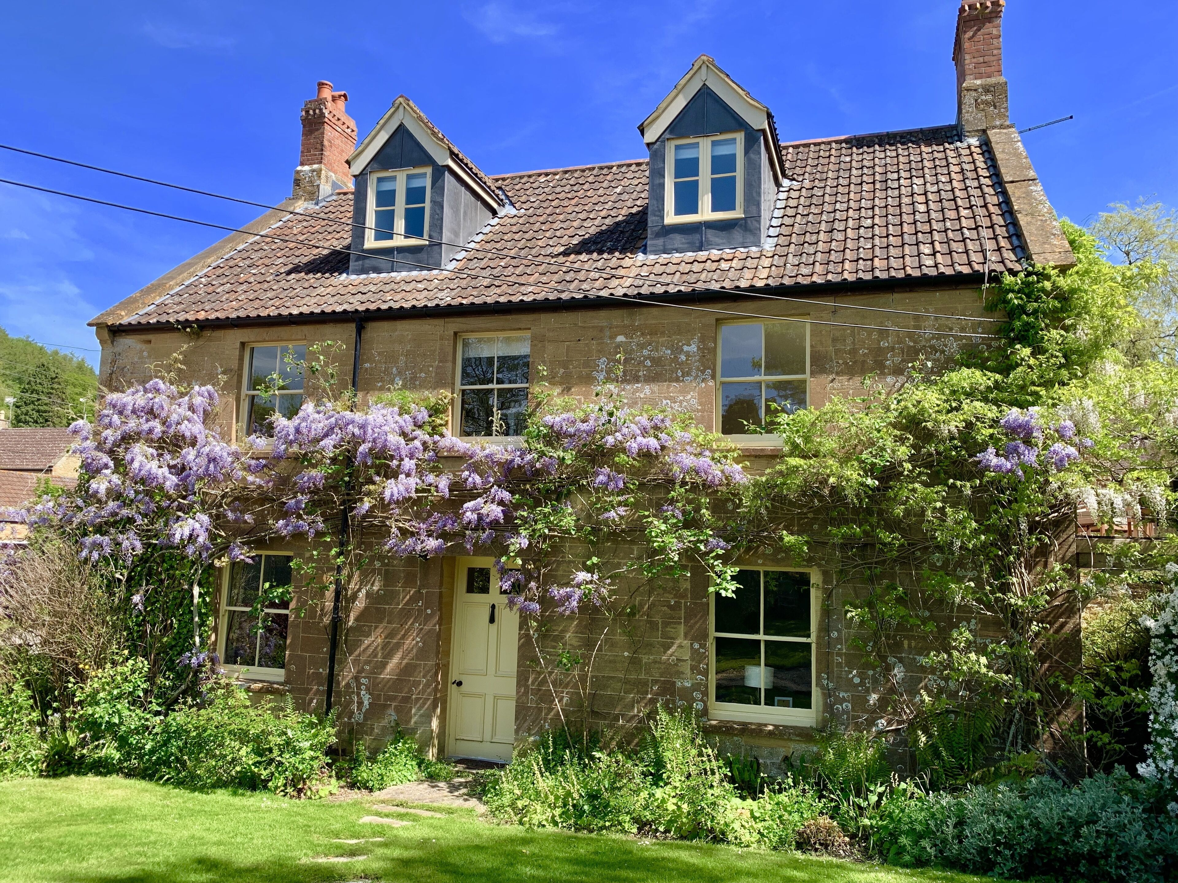 A two-story stone house with a traditional pitched roof, a front door, and a small garden with purple flowering plants.