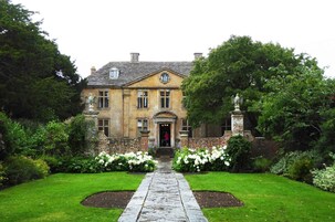 Un edificio histórico con una entrada central, rodeado de jardines cuidados y un camino de piedra que lleva a la puerta.