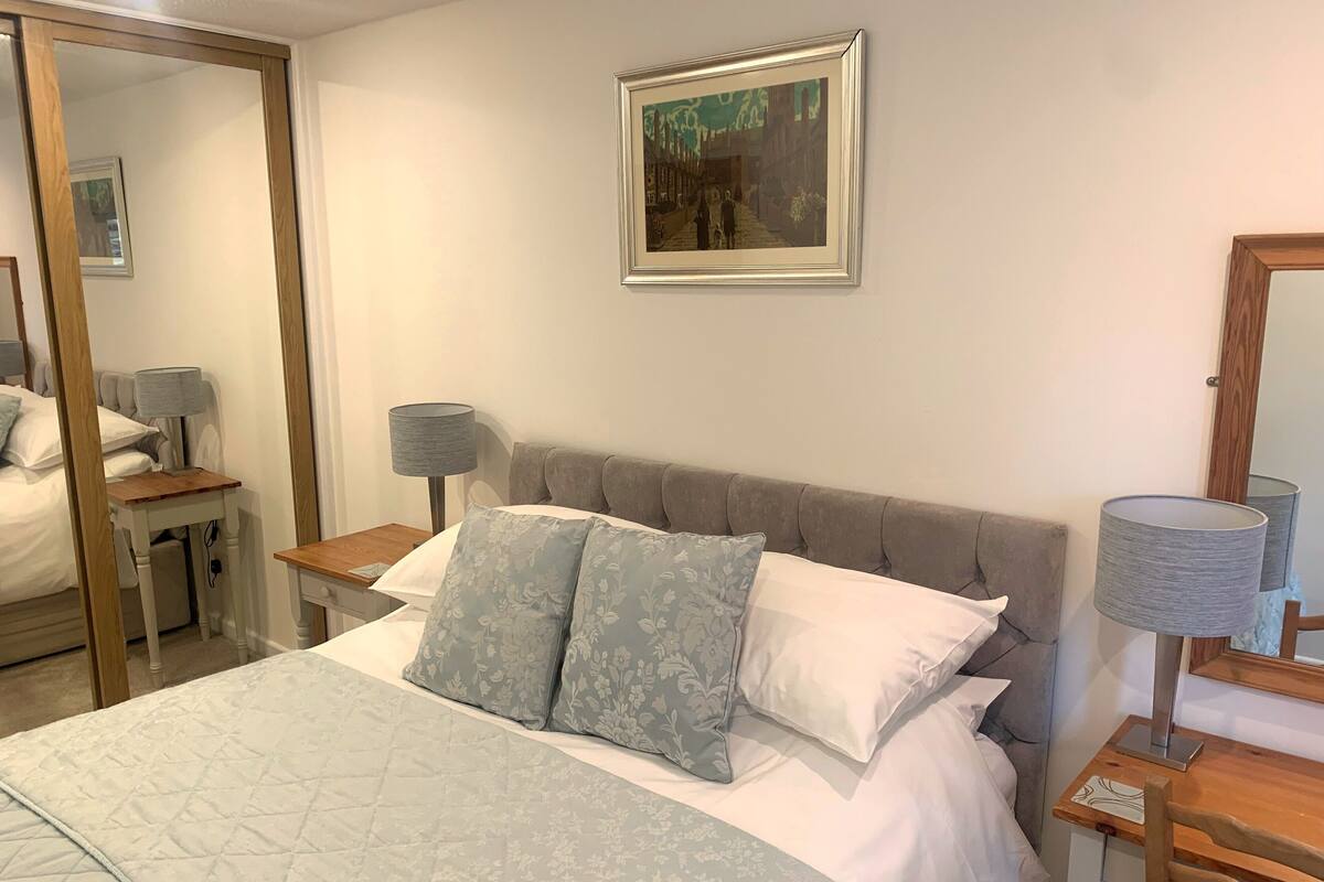 A neatly made bed with a quilted cover, two pillows, a bedside table with a lamp, and a framed picture on the wall.
