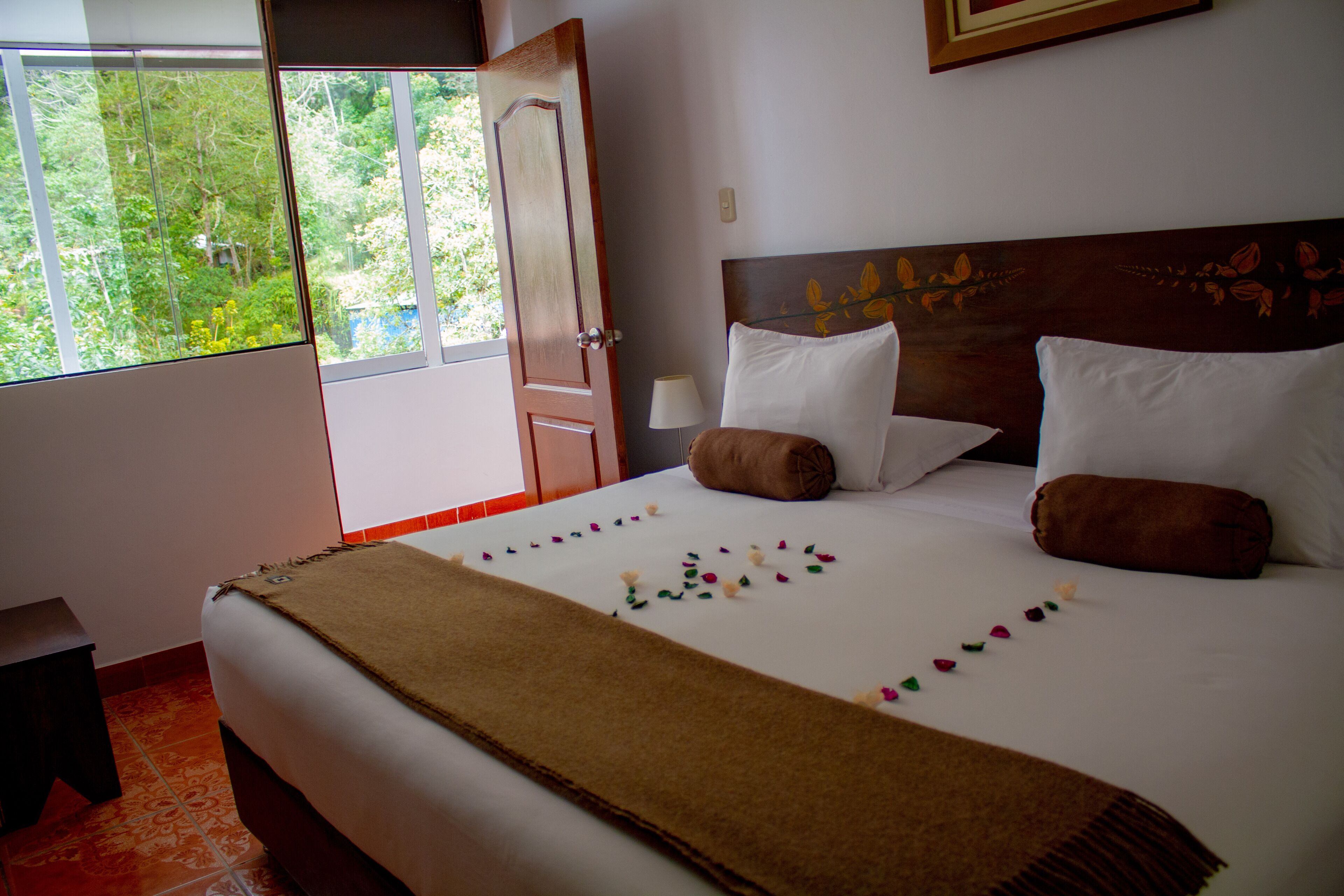 A neatly made bed with a brown throw, white pillows, and a decorative pillow with a leaf pattern. A window with a view of greenery is visible, and a door is open to the outside.