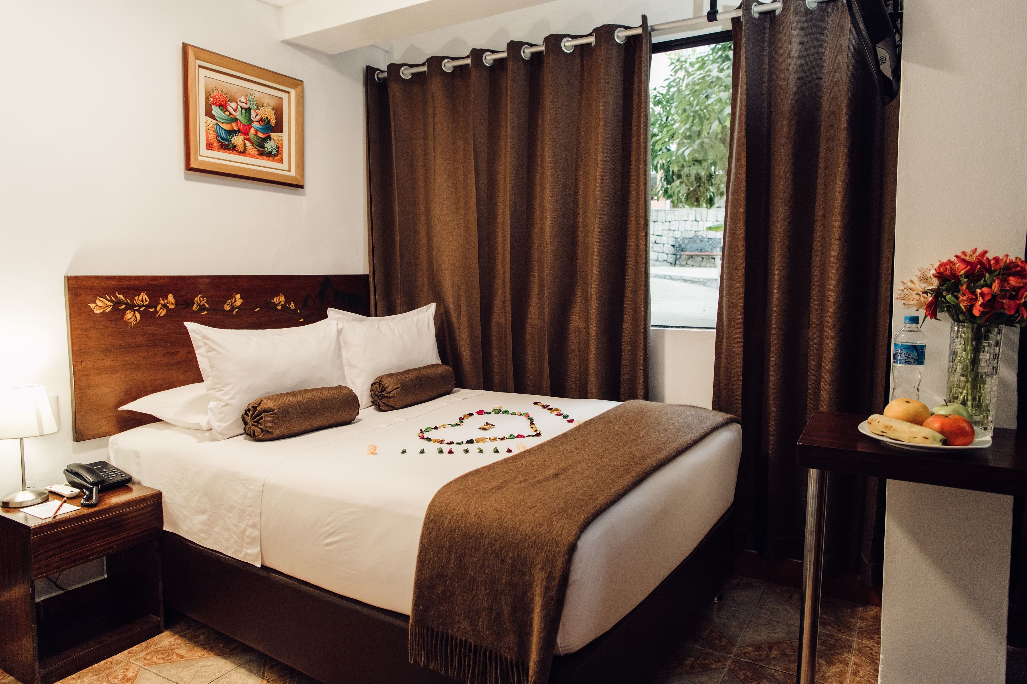 Una camera d'albergo con un letto, un comodino con il telefono, una panca notte con un vaso di fiori e una finestra con tende.