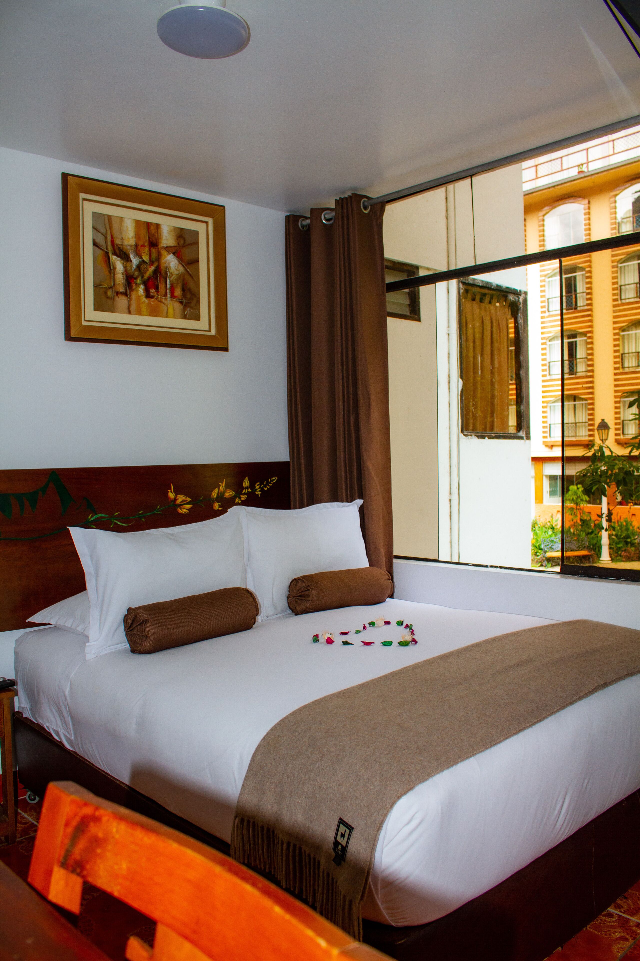 A hotel room with a large bed, a wooden headboard, a painting on the wall, and a view of buildings through the window.