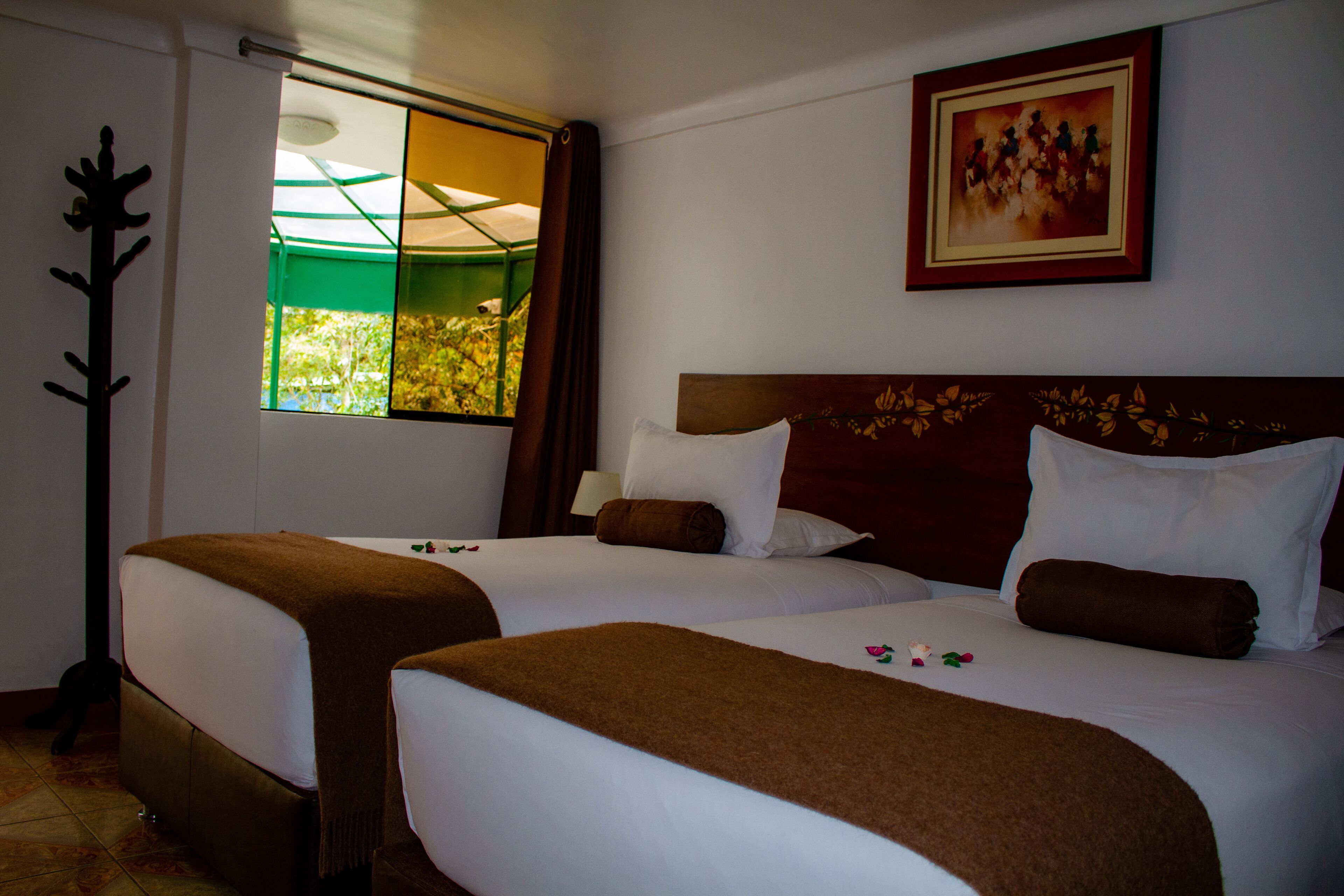 A hotel room with two beds, a window with a view of greenery, a painting on the wall, and a coat rack.