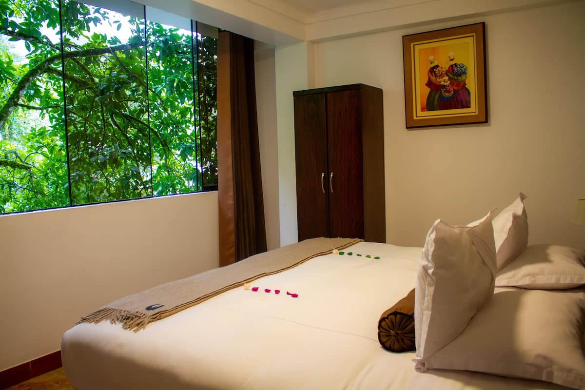 A bedroom with a large window, a bed with white linens, a wooden wardrobe, and a framed picture on the wall.