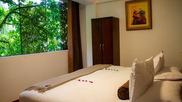 A bedroom with a large window, a bed with white linens, a wooden wardrobe, and a framed picture on the wall.