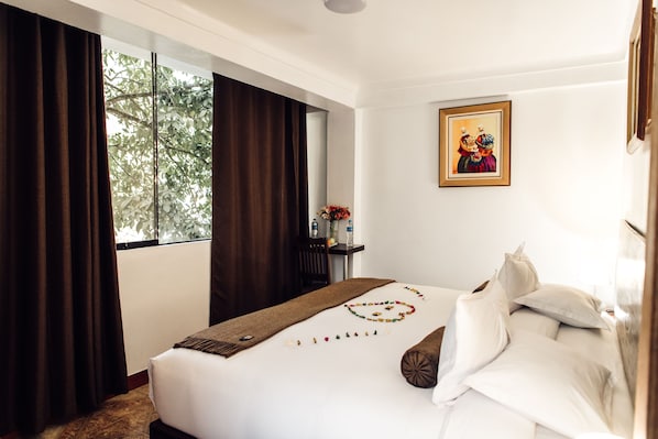 Una habitación de hotel con una cama grande, una ventana con cortinas, un cuadro en la pared y una mesita con un jarrón de flores.