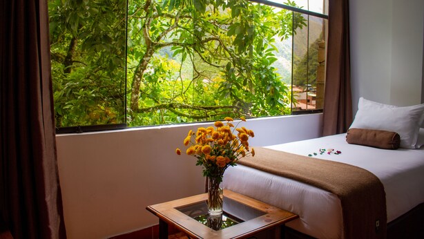 A hotel room with a large window, a bed, a bedside table with a vase of flowers, and a view of greenery outside. - Hotel Retama Machupicchu (Machu Picchu)