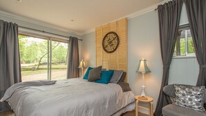 Ein Schlafzimmer mit einem großen Bett, zwei Nachttischen mit Lampen, einem Holz-Kopfteil und Blick auf Grünfläche.