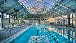 An indoor swimming pool with clear glass walls, lounge chairs, and potted plants.