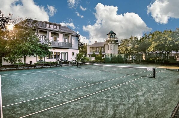 Un campo da tennis con una rete, un edificio con un balcone e una struttura simile a un faro sullo sfondo.
