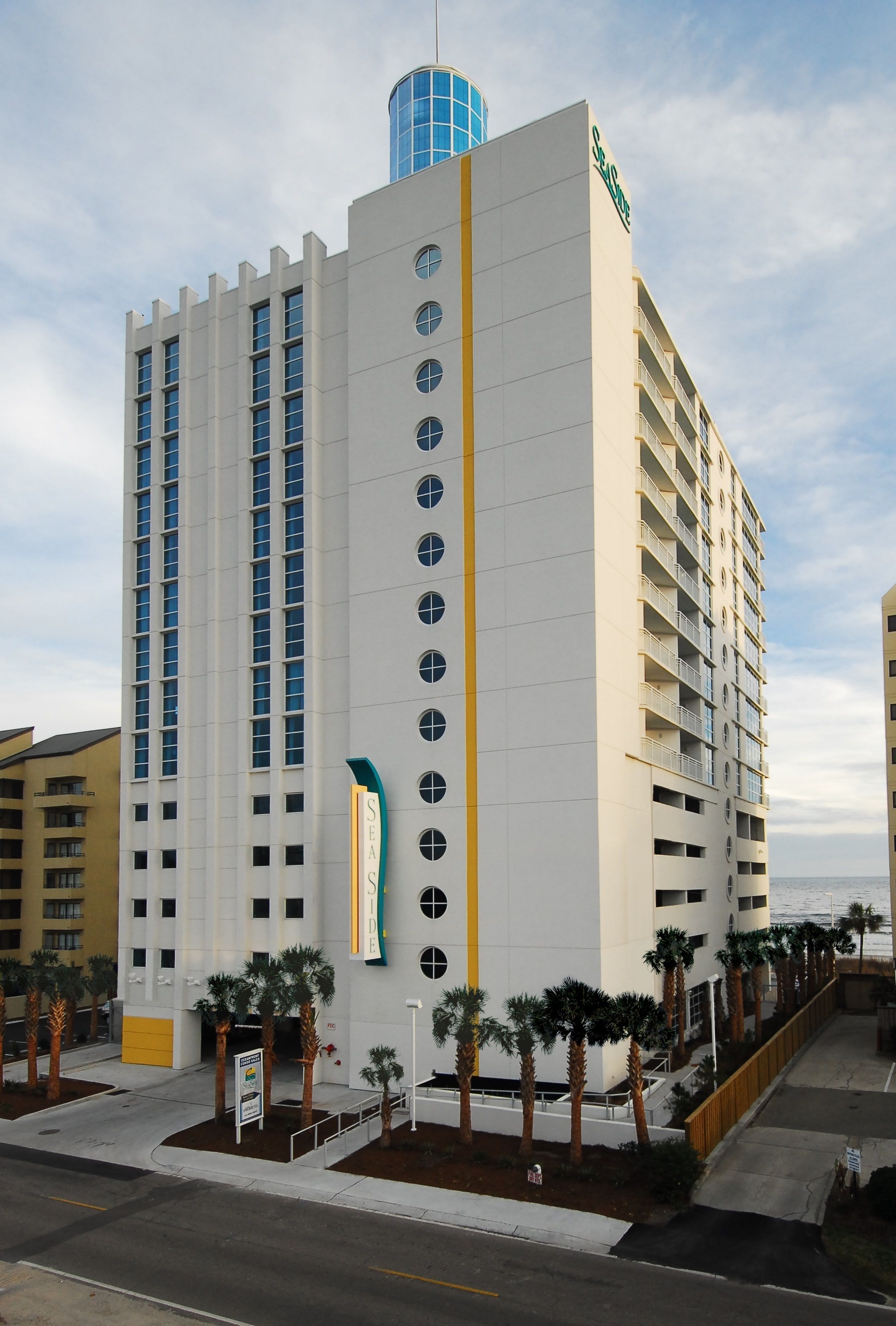 Um hotel alto com design moderno, apresentando uma estrutura cilíndrica azul destacada no topo e uma placa com a inscrição "SEASIDE".