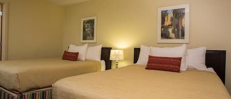 A hotel room with two beds, a striped ottoman, a lamp, and two framed pictures on the wall.