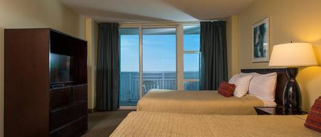 A hotel room with a bed, a TV, a lamp, and a view of the sea through a large window.