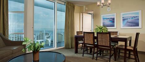 Una habitación de hotel con balcón, una zona de comedor con mesa y sillas, una mesa redonda de centro con una planta y dos cuadros en la pared.