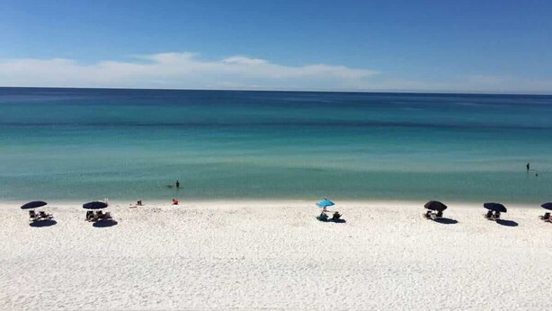 Een strand met wit zand en helderblauw water. Er staan parasols en ligbedden langs de kust.