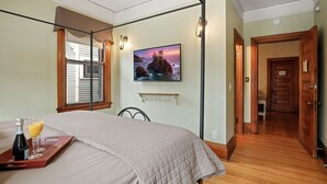 Un dormitorio con una cama, una mesita de noche con una botella y vasos, un cuadro en la pared y suelo de madera.